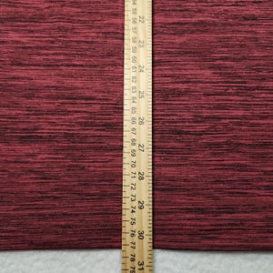 Puede incluir: Primer plano de una tela granate texturizada con motas negras. Una regla de madera con marcas en cent&iacute;metros se coloca verticalmente a lo largo del lado derecho de la tela. La regla mide de 55,88 a 78,74 cm.