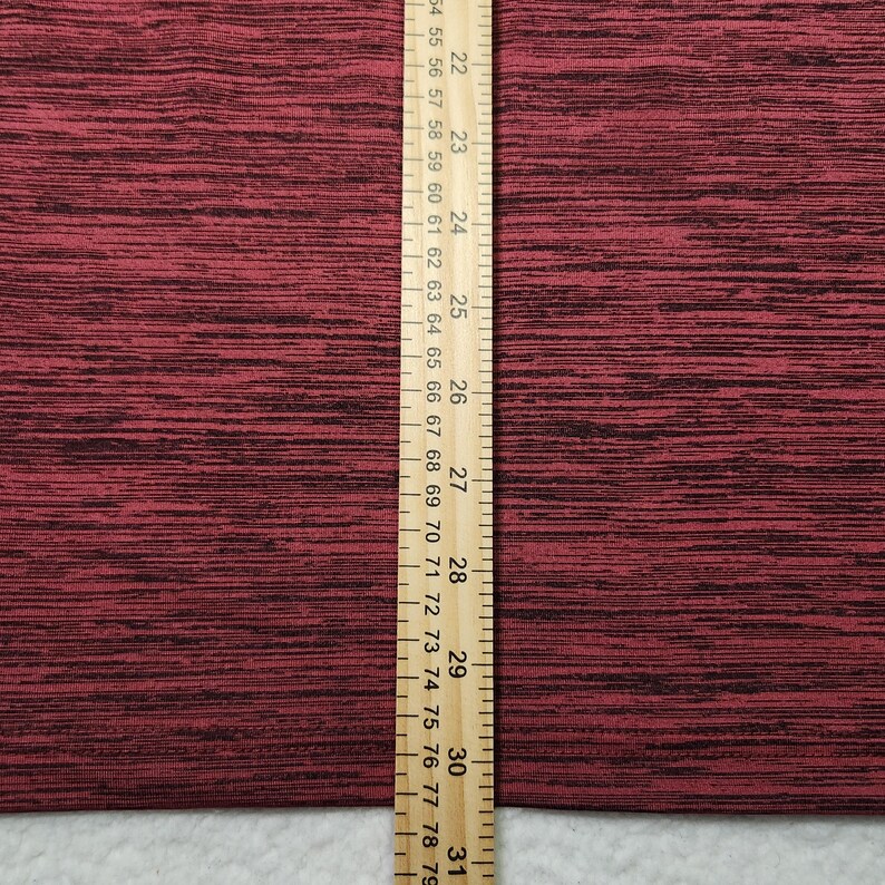 Puede incluir: Primer plano de una tela granate texturizada con motas negras. Una regla de madera con marcas en cent&iacute;metros se coloca verticalmente a lo largo del lado derecho de la tela. La regla mide de 55,88 a 78,74 cm.