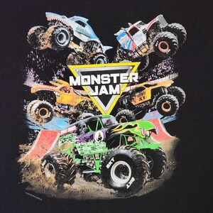 Puede incluir: Camiseta negra con un dise&ntilde;o de varios monster trucks en acci&oacute;n, incluido el Grave Digger. El dise&ntilde;o presenta el logotipo de Monster Jam en un tri&aacute;ngulo amarillo, con camiones de varios colores.