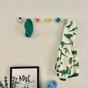 May include: A white wall with a black framed print that says "ADVENTURE" with two arrows crossing each other. There are five wooden hooks on the wall with a green hat, a rocket, a star, a globe, and an astronaut. A blue stuffed elephant is on a wooden table with a green and white dinosaur print jacket hanging on the hook.