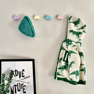 May include: A white and green hooded jacket with a dinosaur print hangs on a wall with pastel cloud-shaped hooks. A green knitted hat hangs on a hook. A black framed print with the text "ADVENTURE" is on a shelf below the hooks. A grey knitted toy is on the shelf.