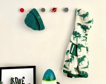 May include: A child's room scene with a white wall adorned with planet-shaped wall hooks. A green beanie and a dinosaur-patterned jacket hang from the hooks. A framed "Adventure" print, a rocket ship toy, and an astronaut figurine are on a wooden surface.