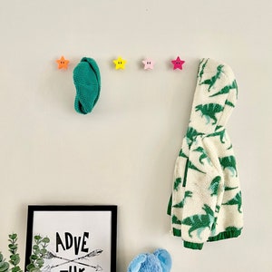 May include: A white wall with five colorful star-shaped hooks. A green hooded sweatshirt hangs on one hook. A blue stuffed elephant sits on a wooden surface below a black framed print with the text "ADVENTURE" in white.