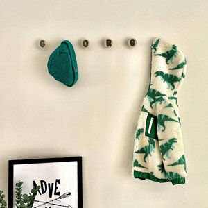May include: A white wall with wooden hooks spelling out the word "GORG" with a green knitted hat hanging on the first hook and a white and green dinosaur print hooded jacket hanging on the last hook. A black framed print with the words "ADVENTURE" and two arrows is on a wooden shelf with a green alien plush toy.