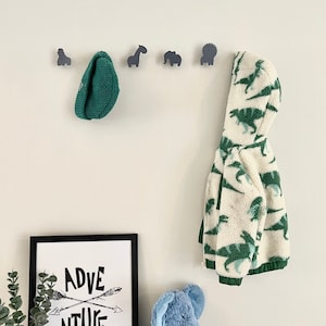 May include: A black framed print with the text "ADVENTURE" and two crossed arrows. A green knitted hat hangs on a wall hook with a grey animal silhouette. A white jacket with a green dinosaur print hangs on another hook. A blue plush elephant sits on a wooden table.