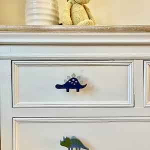 May include: White dresser with two drawers featuring dinosaur-shaped knobs. The top drawer has a blue dinosaur knob, and the bottom drawer has a green dinosaur knob.