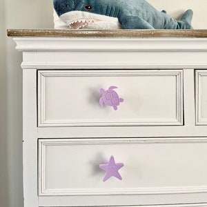 May include: A white dresser with two drawers. The top drawer has a purple turtle-shaped knob and the bottom drawer has a purple starfish-shaped knob.
