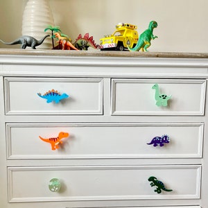 May include: White chest of drawers with six drawers featuring dinosaur-shaped drawer pulls. The pulls are in various colours, including blue, green, orange, purple, and green.