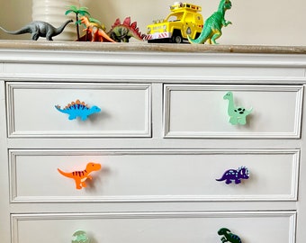 May include: White dresser with six drawers featuring dinosaur-shaped drawer pulls. The pulls are in various colors, including blue, green, orange, purple, and green.