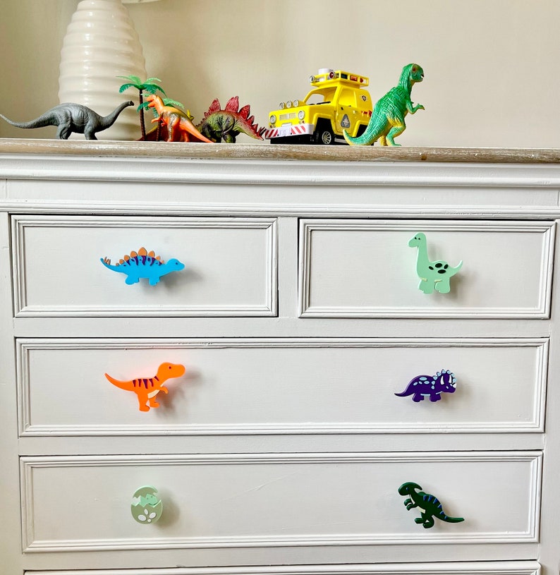 May include: White chest of drawers with six drawers featuring dinosaur-shaped drawer pulls. The pulls are in various colours, including blue, green, orange, purple, and green.