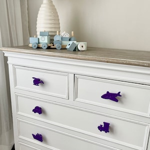 May include: White chest of drawers with four drawers and purple toy-shaped drawer pulls. The pulls are a helicopter, a car, a ship, and an aeroplane. A toy train is on top of the dresser.