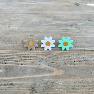 Op de afbeelding: Drie kleine daisy-vormige bloemdecoraties in verschillende kleuren. De bloemen zijn bruin, wit en groen met een gouden midden.
