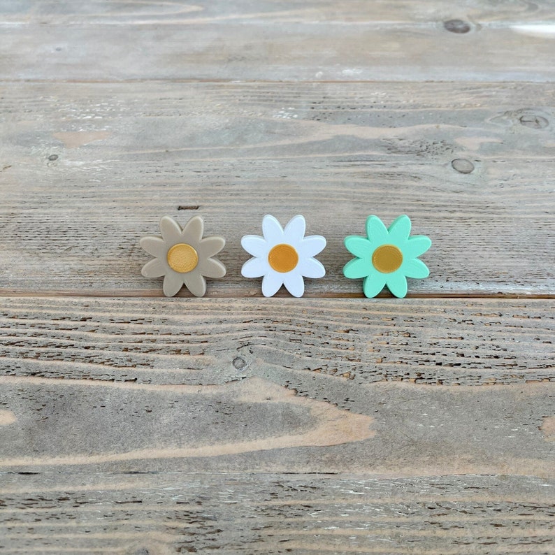 Op de afbeelding: Drie kleine daisy-vormige bloemdecoraties in verschillende kleuren. De bloemen zijn bruin, wit en groen met een gouden midden.