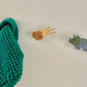 Peut inclure: Un tissu tricot&eacute; vert est accroch&eacute; &agrave; un mur blanc &agrave; c&ocirc;t&eacute; d'un crochet mural en forme de dinosaure brun et or et d'un crochet mural en forme de dinosaure bleu et vert.