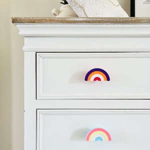 May include: White chest of drawers with two drawers. The top drawer has a rainbow-coloured drawer pull with purple, orange, red, and white stripes. The bottom drawer has a rainbow-coloured drawer pull with pink, yellow, blue, and white stripes.