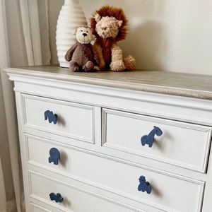 May include: White dresser with four drawers and blue animal-shaped drawer pulls. The top drawer has an elephant pull, the second drawer has a giraffe pull, the third drawer has a lion pull, and the bottom drawer has a hippo pull.