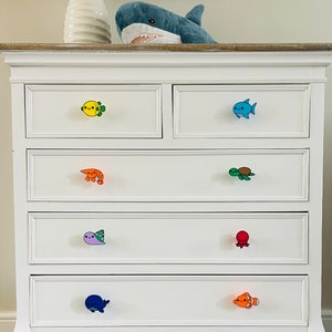 May include: White chest of drawers with marine life-themed drawer knobs. The knobs feature a yellow fish, blue fish, orange shrimp, green turtle, purple sea creature, red octopus, blue whale, and orange clownfish. A shark soft toy sits on top.
