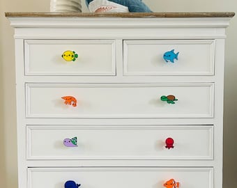 May include: White dresser with colorful sea creature drawer pulls. The pulls include a yellow fish, blue fish, orange shrimp, green turtle, purple sea creature, red octopus, blue whale, and orange clownfish. A plush shark toy sits on top.