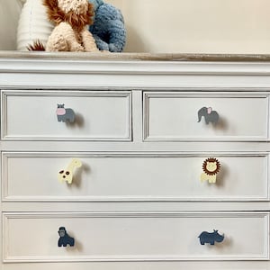 May include: White chest of drawers with six drawers, each with a different animal-shaped knob. The knobs are a grey hippopotamus, a grey elephant, a yellow giraffe, a yellow lion, a grey gorilla, and a grey rhinoceros.