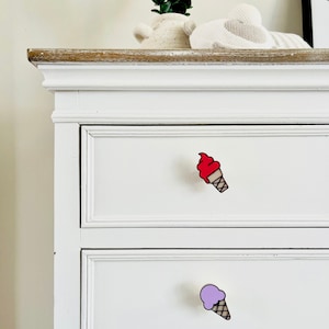 May include: White dresser with two drawers. The top drawer has a red ice cream cone pull and the bottom drawer has a purple ice cream cone pull.