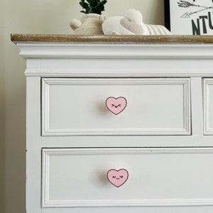 May include: White dresser with two drawers. Each drawer has a pink heart-shaped knob with a smiling face.