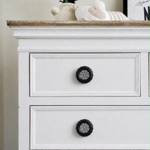 Puede incluir: Una cómoda de madera blanca con dos cajones que tienen pomos en forma de neumático negro y plateado. La cómoda presenta un borde superior en tono madera natural y un diseño de marco rectangular en los frentes de los cajones.