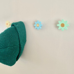 Op de afbeelding: Drie bloemvormige haken aan een witte muur, met een groene gebreide muts die aan een van de haken hangt. De haken zijn geschilderd in tinten blauw en groen, met gele centra.