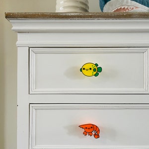 May include: White chest of drawers with two drawers. The top drawer has a yellow and green pufferfish drawer pull. The bottom drawer has an orange and red shrimp drawer pull.