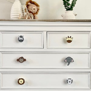 May include: White chest of drawers with six drawers, each featuring a different animal-themed knob. The knobs are a zebra, giraffe, monkey, elephant, lion, and a zebra.