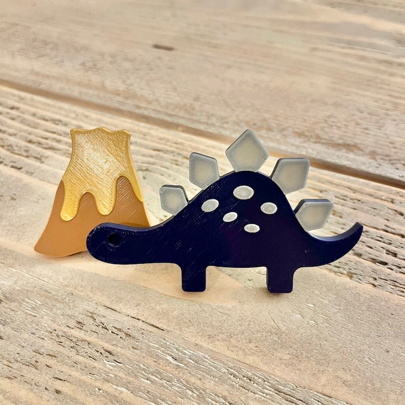 Peut inclure: Un jouet de dinosaure en bois bleu et gris avec un jouet de volcan en bois jaune et brun. Le dinosaure a un dos h&eacute;riss&eacute; et des points blancs.