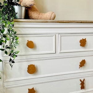 May include: White chest of drawers with six drawers featuring wooden knobs shaped like woodland animals, including an acorn, squirrel, owl, leaf, rabbit, and fox. The knobs are painted gold.