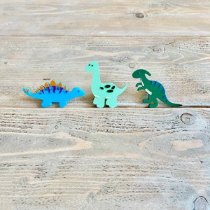 May include: Three colourful dinosaur figurines on a wooden surface. The dinosaurs are green, blue, and a light green colour.