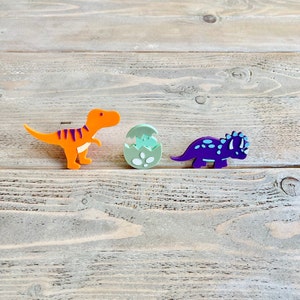 May include: Three colourful dinosaur figurines on a wooden surface. An orange dinosaur, a green dinosaur egg, and a purple dinosaur.