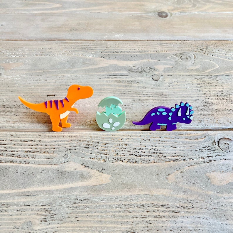 May include: Three colourful dinosaur figurines on a wooden surface. An orange dinosaur, a green dinosaur egg, and a purple dinosaur.