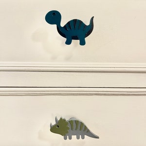 May include: Two wooden dinosaur drawer pulls. The top pull is a blue dinosaur with black stripes. The bottom pull is a green and gray dinosaur with black stripes.