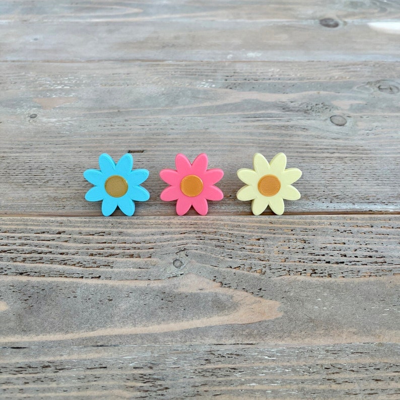 Op de afbeelding: Drie kleurrijke bloemvormige knopen in blauw, roze en geel. De knopen zijn in een rij op een houten oppervlak gerangschikt.