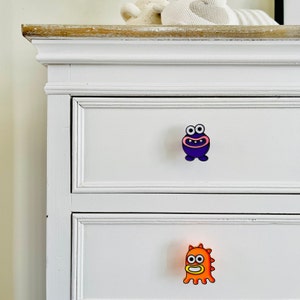 May include: White dresser with two drawers. The top drawer has a purple monster-shaped knob. The bottom drawer has an orange monster-shaped knob.