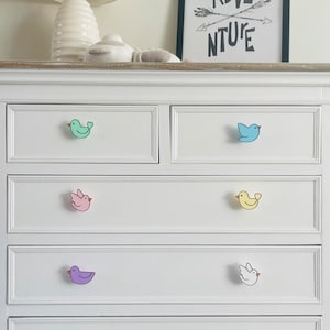 May include: White chest of drawers with six drawers, each with a different coloured bird-shaped knob. The knobs are green, pink, yellow, white, and purple.