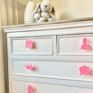 May include: White chest of drawers with six drawers featuring pink plastic drawer pulls shaped like a castle, crown, carriage, frog, and mirror.