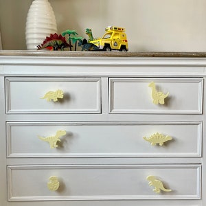 May include: A white dresser with four drawers. Each drawer has a yellow dinosaur-shaped pull. The top drawer has a yellow dinosaur with a long neck, the second drawer has a yellow dinosaur with spikes on its back, the third drawer has a yellow dinosaur with a long tail, and the bottom drawer has a yellow dinosaur with a round head.