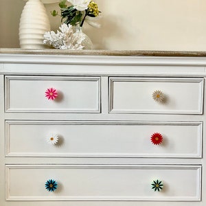 May include: White chest of drawers with six drawers, each with a different coloured flower-shaped knob. The knobs are pink, white, red, blue, and green.