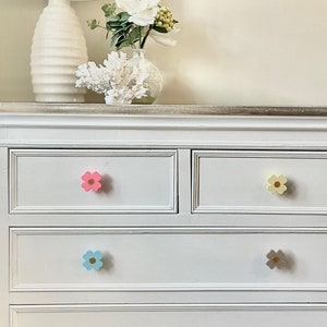 May include: White chest of drawers with four drawers and flower-shaped knobs. The knobs are pink, yellow, blue and grey.