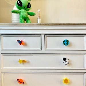 May include: White chest of drawers with five drawers featuring space-themed drawer pulls. The pulls include a rocket, a planet, a shooting star, an astronaut, and a sun.