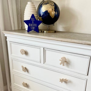 May include: White chest of drawers with four drawers and wooden knobs. The knobs are shaped like a seashell, a butterfly, a star, and an astronaut. The dresser has a light wood top and a distressed white finish.