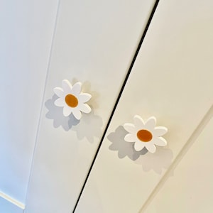 May include: Two white cabinet knobs shaped like daisies with yellow centres. The knobs are mounted on a white cabinet door.