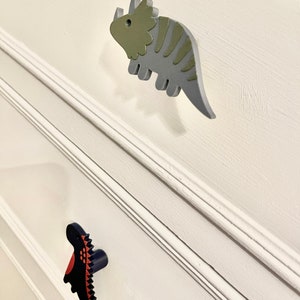 May include: Two dinosaur-shaped drawer pulls. One is a green and gray triceratops, the other is a black and red dinosaur.