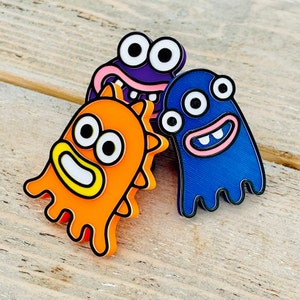 May include: Three monster-shaped pins in vibrant colours. The pins are orange, purple, and blue, each with a distinct design. The orange monster has spiky edges, the purple one has a wide grin, and the blue one has a wavy bottom edge. All have large eyes and white teeth.