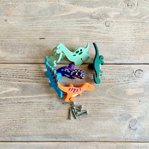 May include: Five colourful dinosaur-shaped drawer pulls, including green, blue, purple, and orange, on a wooden surface.