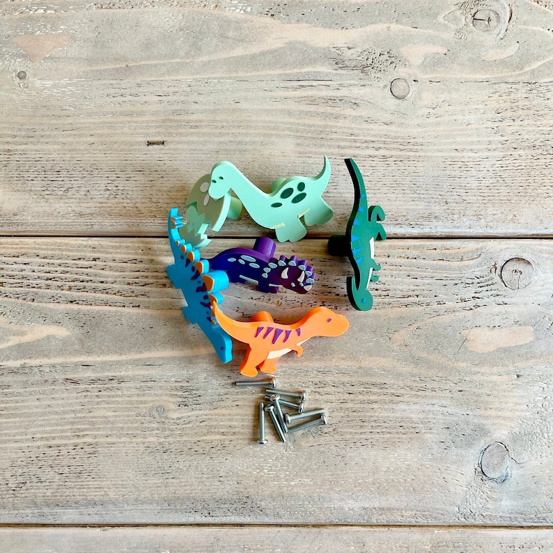 May include: Five colourful dinosaur-shaped drawer pulls, including green, blue, purple, and orange, on a wooden surface.