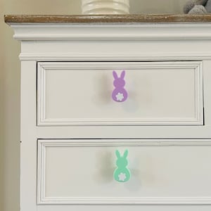 May include: White chest of drawers with two drawers. The top drawer has a purple bunny rabbit pull and the bottom drawer has a green bunny rabbit pull.